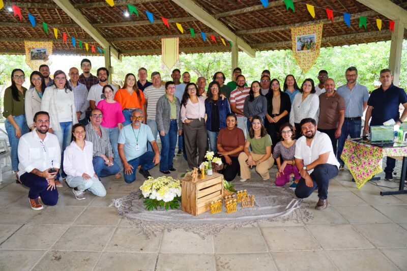 Large diverse group of people posing for a group photo under a decorated outdoor pavilion with colorful bunting and flowers in the center.