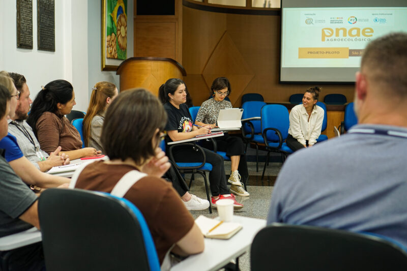 People seated in a circle in a conference room, a speaker uses a laptop while others listen and take notes near a projection screen.